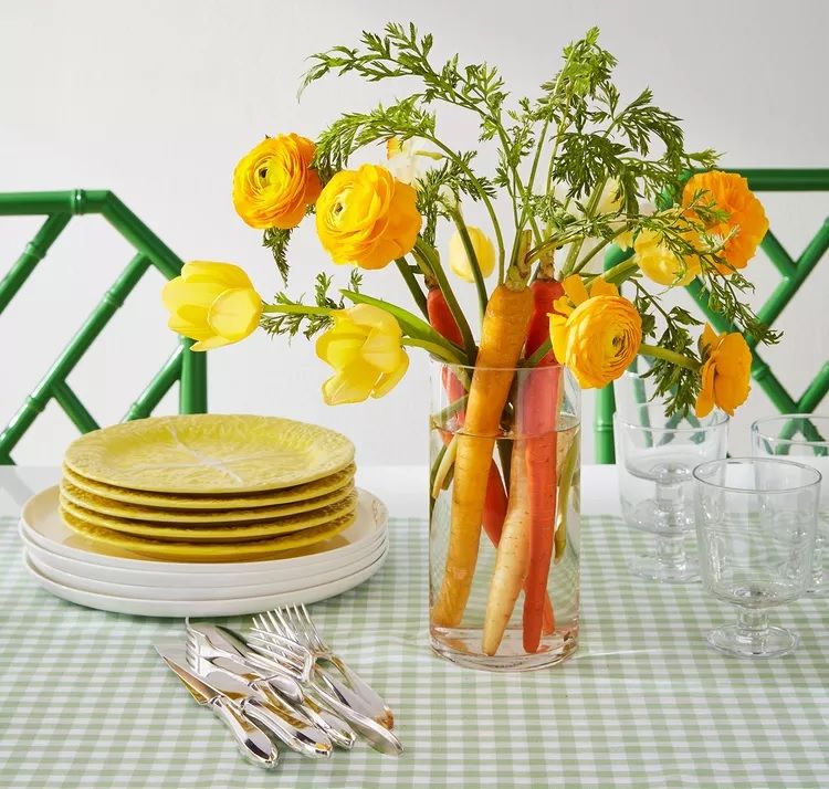 Váza s vodou, žlutými květinami a mrkvemi jako květinová dekorace na velikonoční stůl
