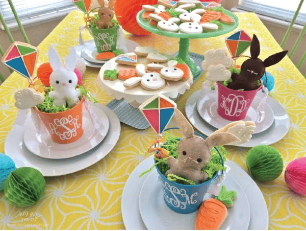 Prostřený stůl s barevnými kyblíčky s plyšovými zajíčky, perníčky a papírovými barevnými ozdobami pro dětskou párty