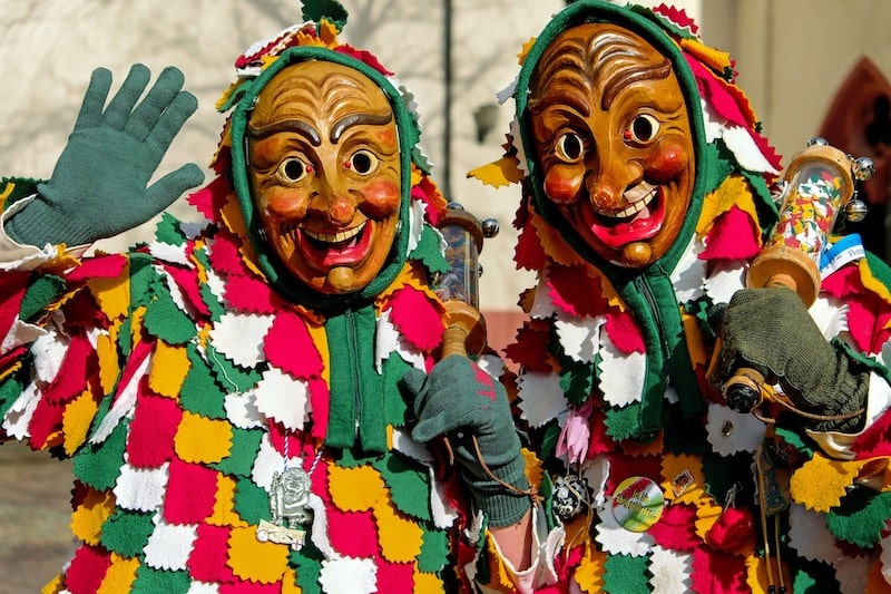 Dvě osoby v barevných šatech a typických dřevěných maskách se usmívají přímo do kamery.